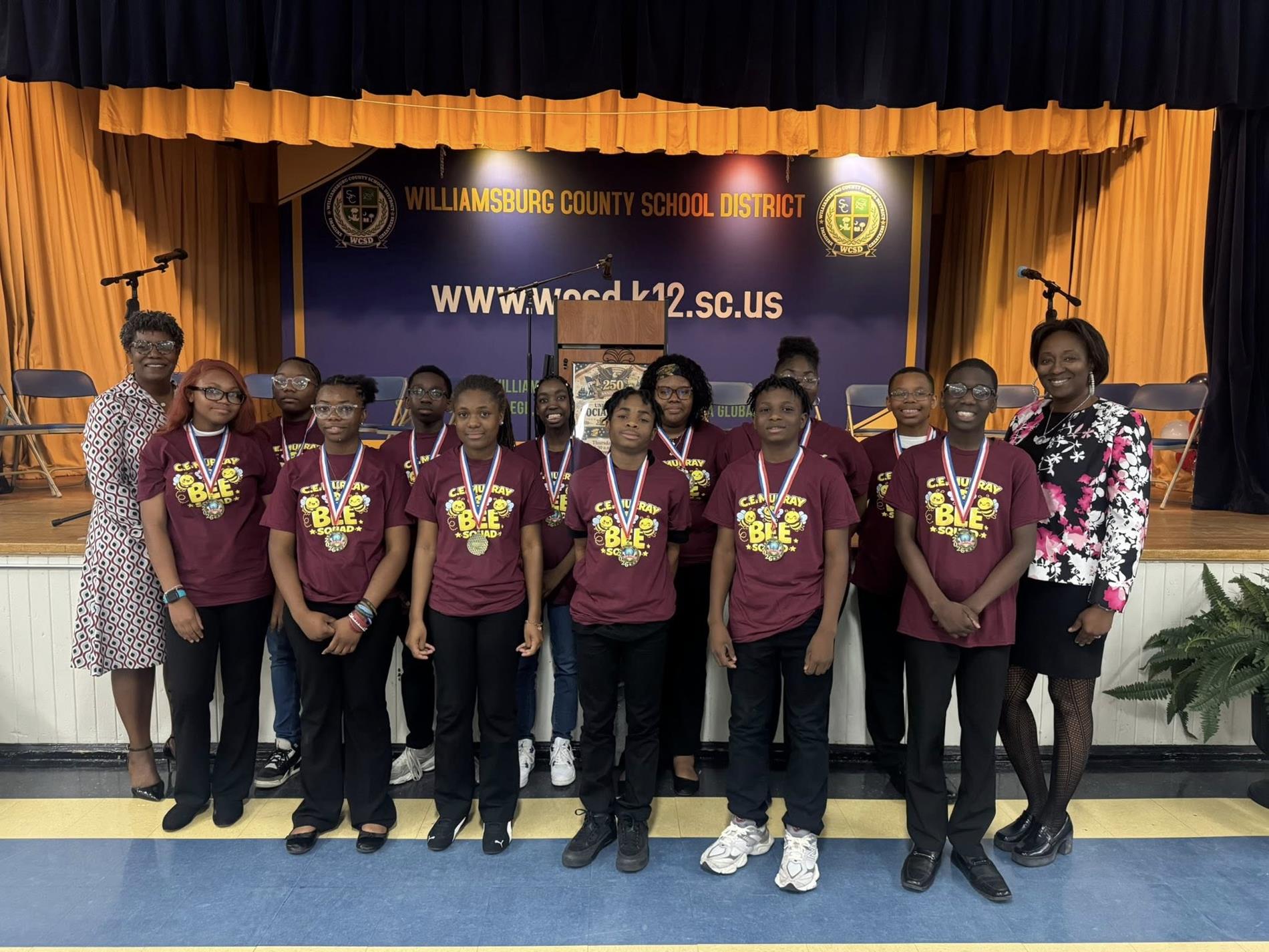 A group of middle school students stand together on a stage in front of a banner reading “Williamsburg County School District” and a website URL. The students wear matching maroon shirts and medals around their necks, posing in two rows. Two adults stand on either side of the group, smiling. The stage has gold curtains, microphones, and a podium in the background, suggesting an awards or recognition event.