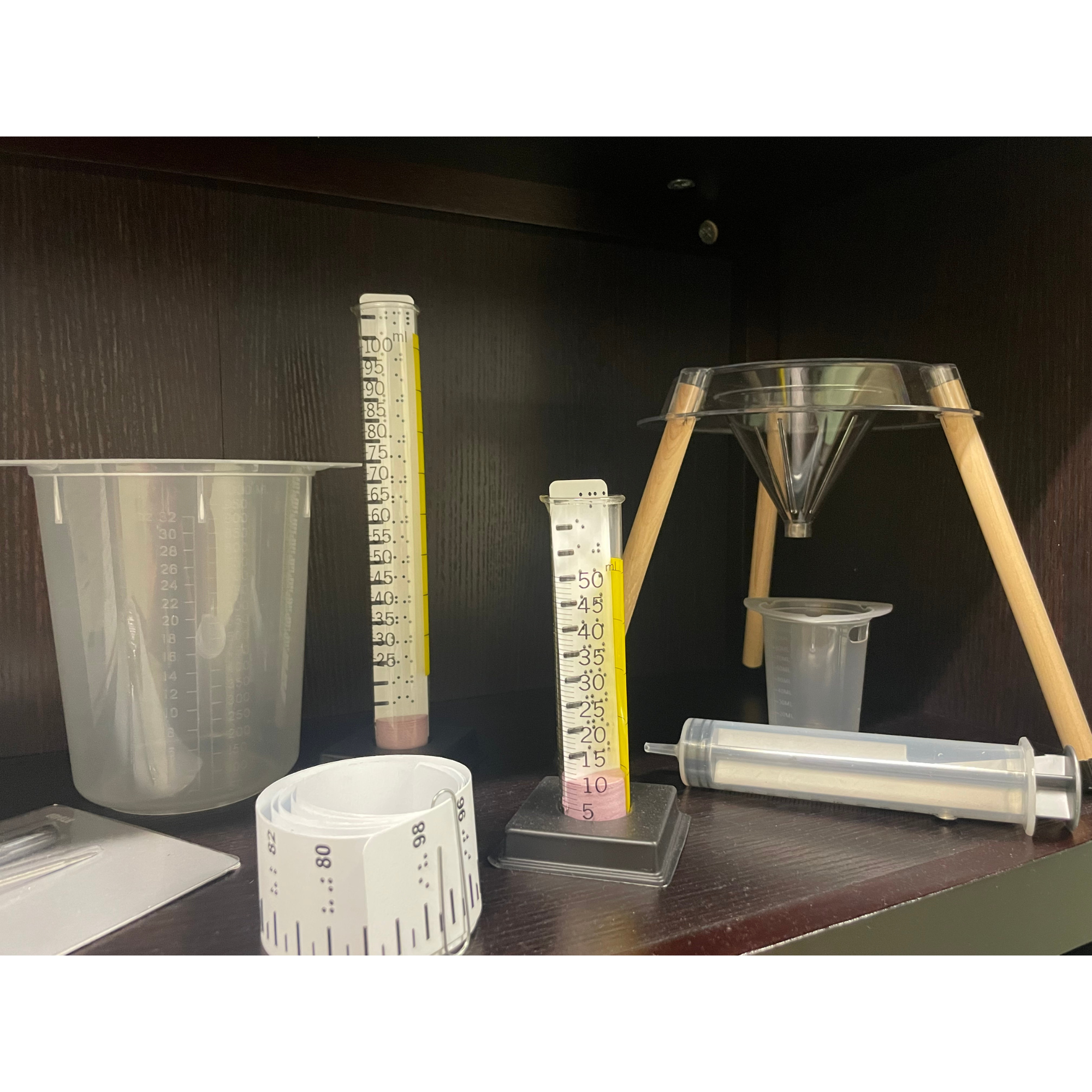 Assorted science lab equipment on a shelf, including measuring cylinders with liquid, a beaker, a funnel on a stand, a syringe, and a measuring tape.