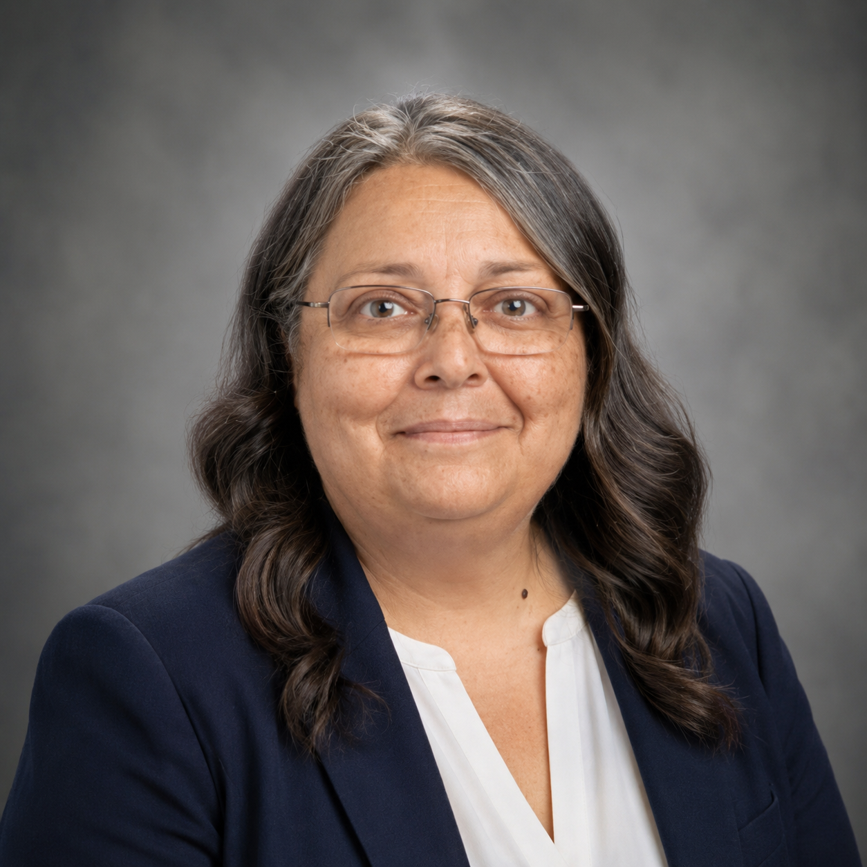 Professional headshot of Nettie Jeansonne smiling gently and facing the camera. She is wearing a navy blazer over a white blouse and rectangular glasses. Her shoulder-length dark hair with gray highlights is styled in loose curls. She is posed against a soft gray studio background, creating a polished portrait suitable for use in an instructional coach or school leadership announcement.