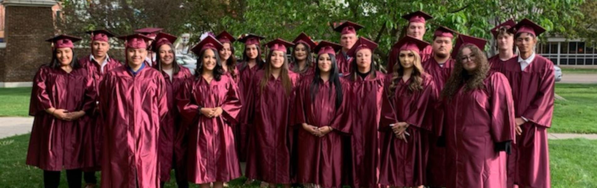 Eighteen graduates in maroon cap and gowns stand under trees in a park. They are standing in a single row.