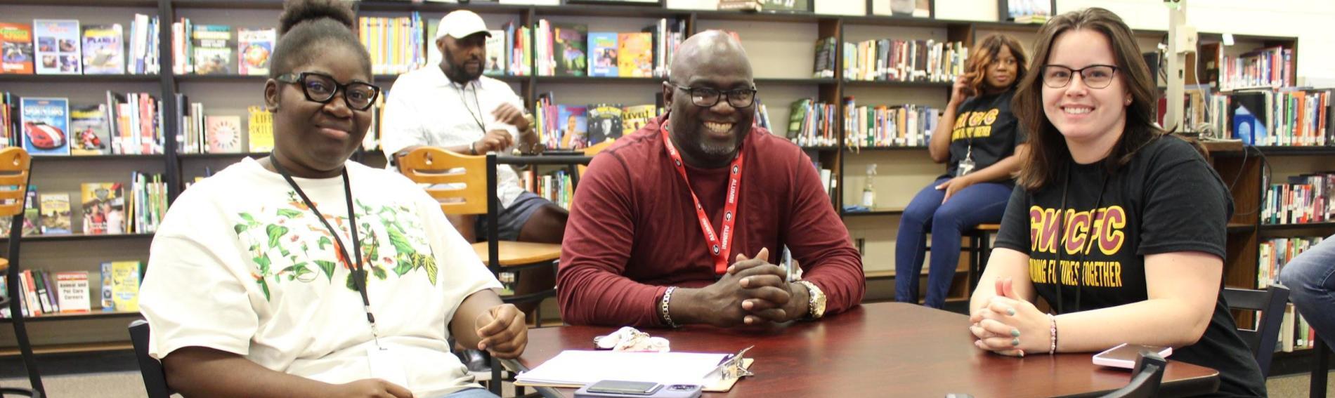 One male teacher and two female teachers smile at the camera while sitting at a table together in the media center