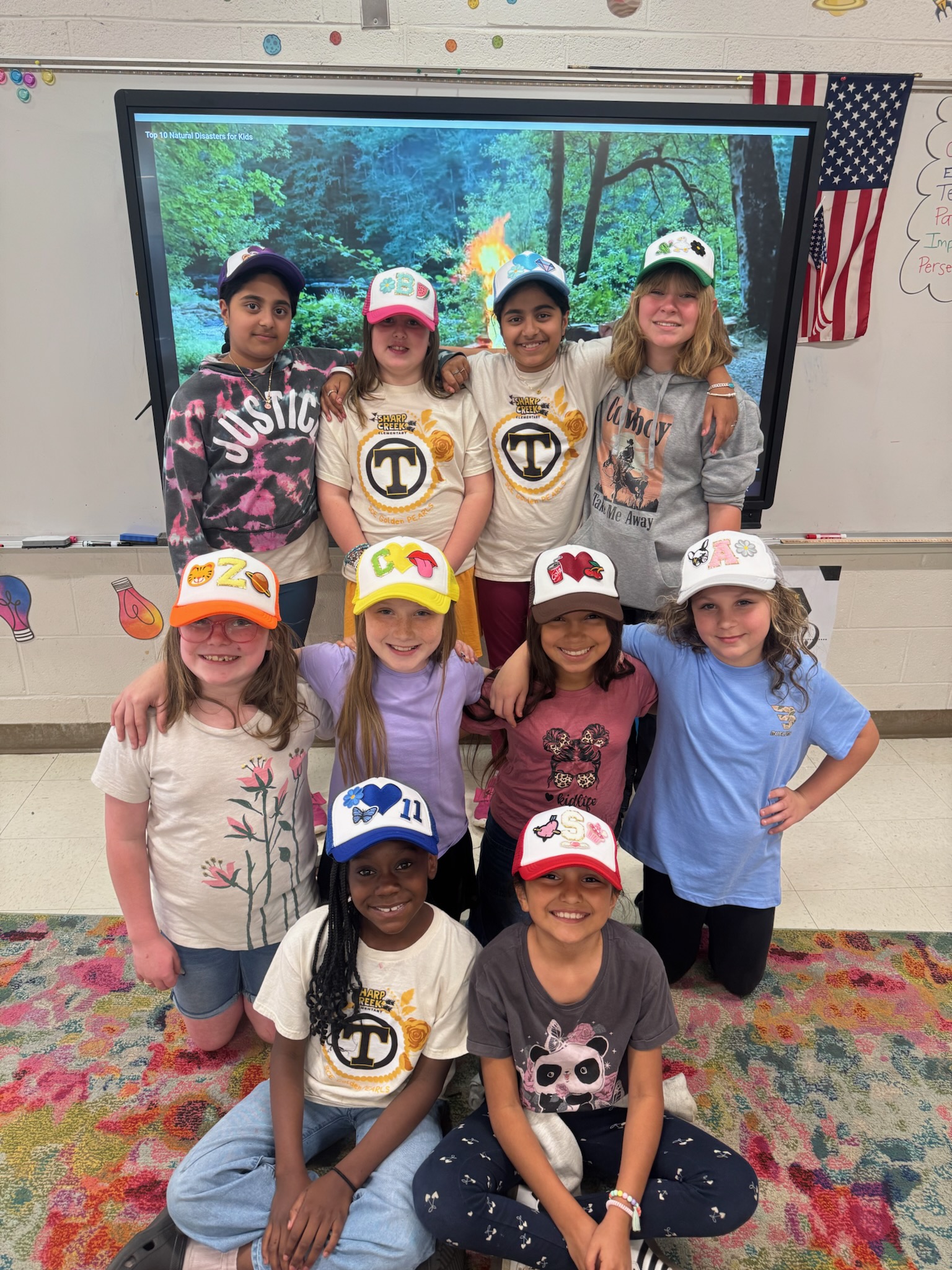 A group of smiling elementary students pose together in a classroom, proudly wearing colorful, decorated baseball caps they created. Students stand, kneel, and sit in front of an interactive display and classroom whiteboard.