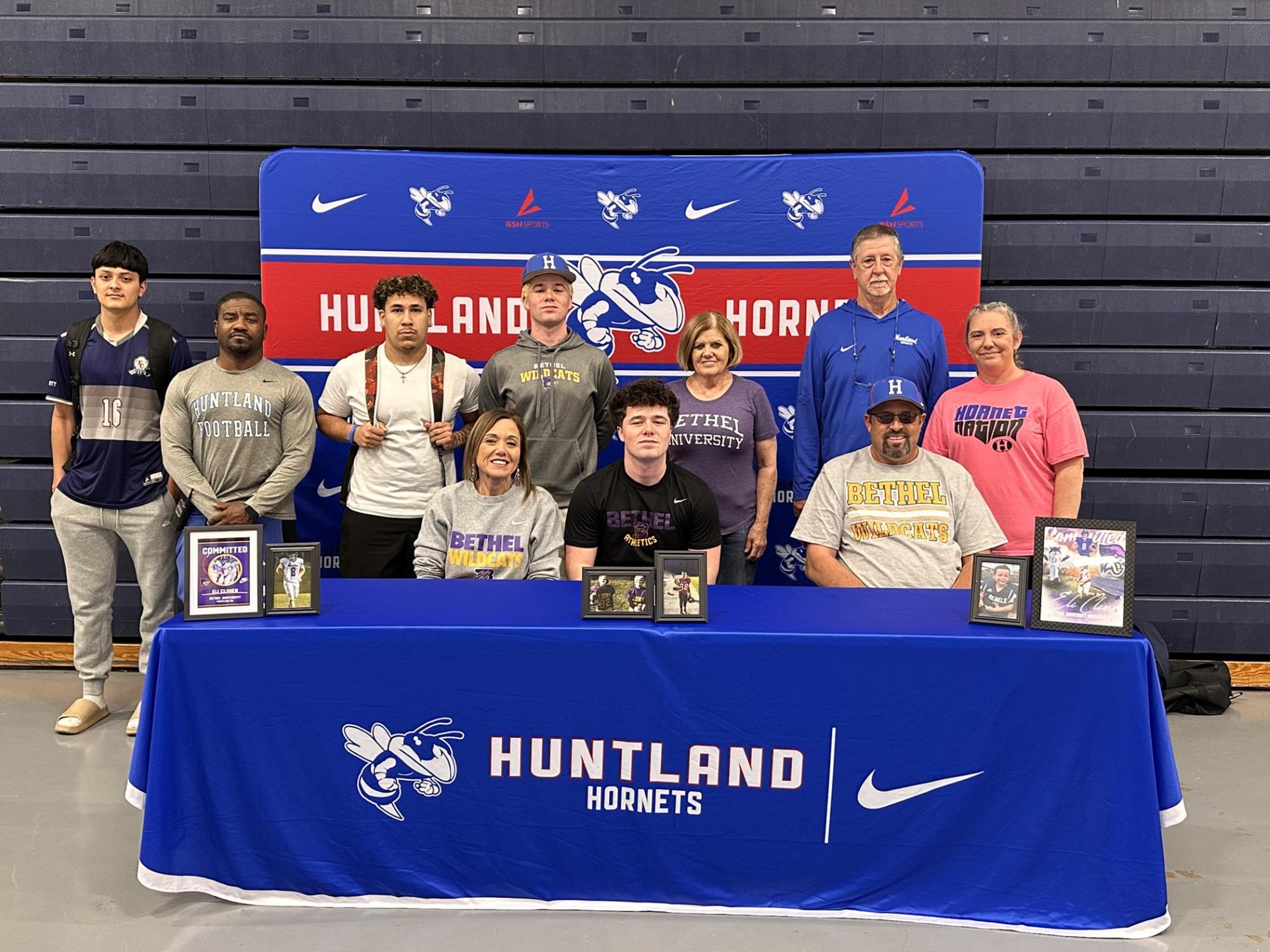 Photo of Eli Climer surrounded by friends, family, and coaches to celebrate signing with Bethel University. Table with photos from Eli's football career on a table with blue table cloth, backdrop blue with hornet logo, bsn sports logo, and nike logo, red lettering that reads, "HUNTLAND HORNETS"  and our mascot image in the middle.
