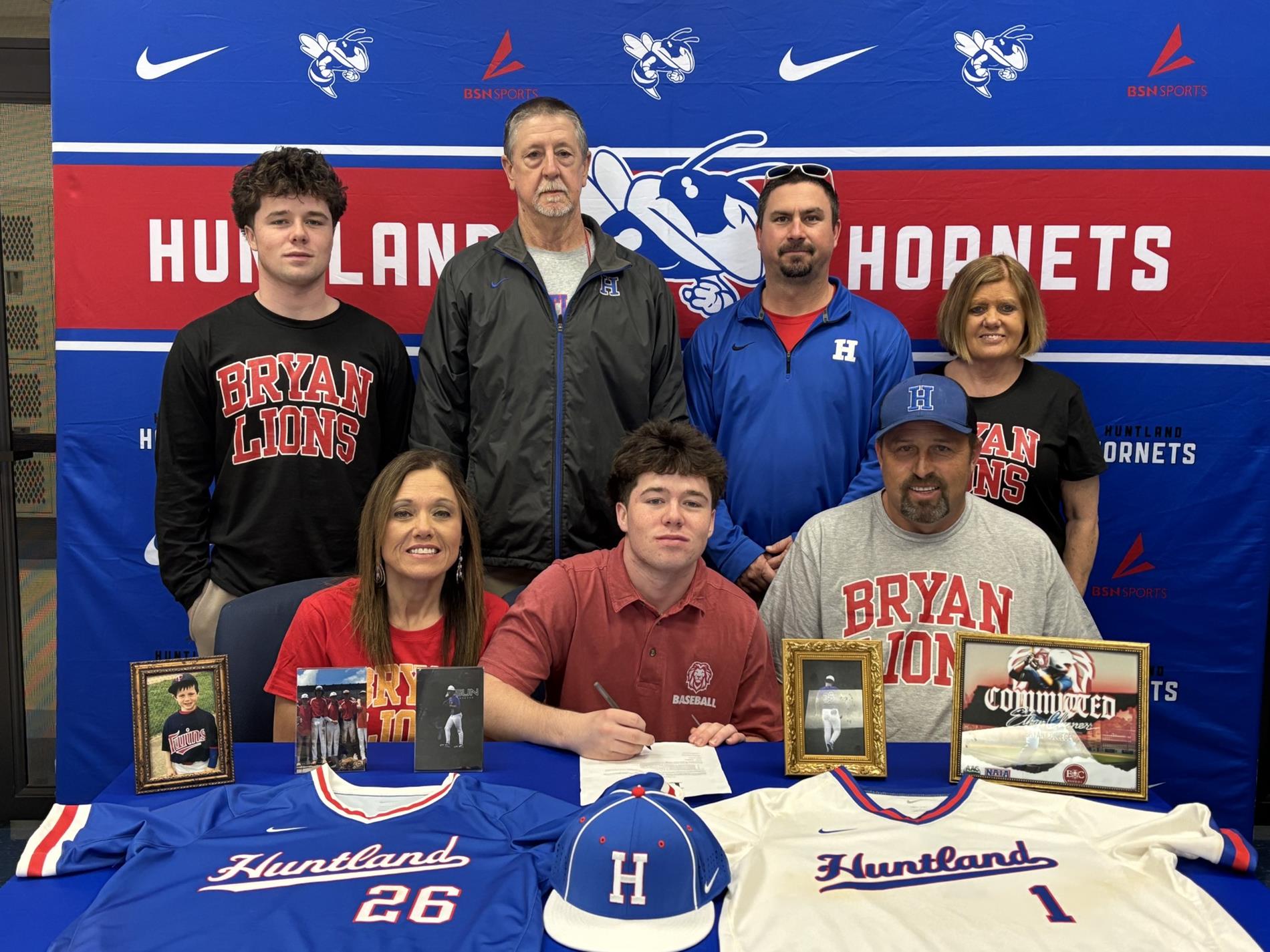 Picture of Ethan Climer signing to continue his baseball career. front left to right, Donna Humphrey, Ethan Climer, Chad Powell, back row- Eli Climer, Coach Rob, Coach Brent, Felicia Sons. on the table are his High school baseball jerseys, hat and baseball photos.